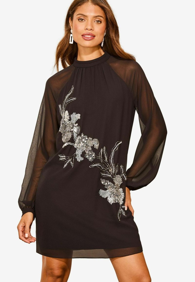 Rochie mini neagră cu mâneci lungi transparente, având broderie florală intricate în nuanțe argintii pe partea din față și pliere subtilă la decolteu.