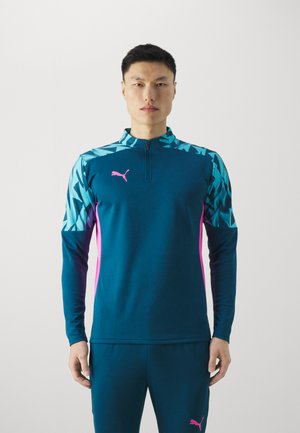 Maglietta sportiva a maniche lunghe color teal con motivi geometrici blu, dettagli rosa e colletto con mezza zip. Presenta un tessuto testurizzato e il logo Puma.