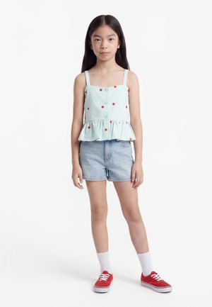 Fată tânără cu păr lung și închis la culoare, purtând un top bleu deschis cu buline, fără mâneci, pantaloni scurți din denim, șosete albe și teniși roșii.