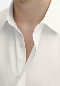 Chemise blanche boutonnée avec un col pointu, présentant une texture de tissu lisse et des boutons visibles, partiellement déboutonnée au niveau du cou.