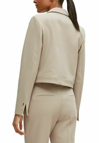 Beige maßgeschneiderte Jacke mit einem cropped Design, Reverskragen und seitlichen Schlitzen an den Ärmeln, gefertigt aus einem strukturierten Stoff.
