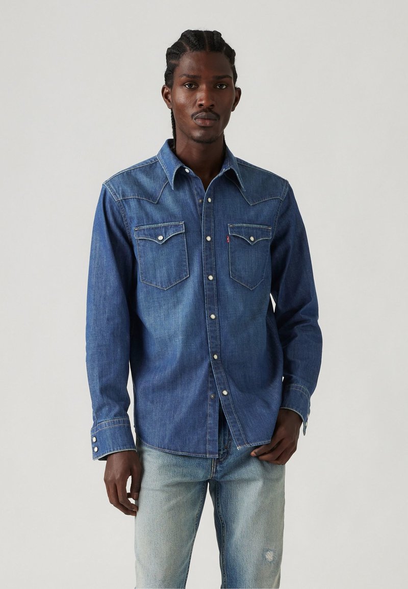 Camisa de mezclilla en azul oscuro con un yugo de estilo occidental, dos bolsillos en el pecho, botones de metal y costuras claras en los puños y el cuello.
