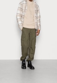 Beige- och vitrutigt skjorta över en beige tröja, ihop med olivgröna cargobyxor och svarta ankelstövlar. Texturerade tyger och praktisk design.