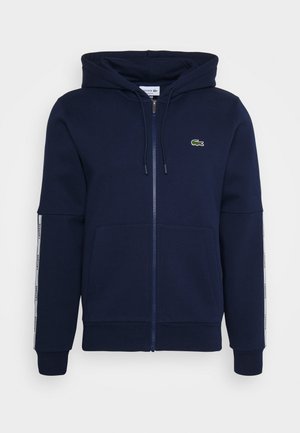Marineblauwe hoodie met ritssluiting, voorzakken, capuchon met trekkoord, witte zijstrepen op de mouwen en een klein groen krokodillenlogo op de borst.