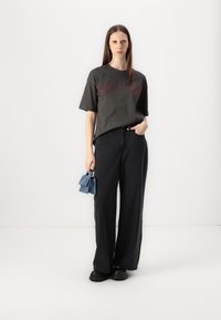 Šedé oversized tričko s červeným grafickým textem, doplněné o široké černé džíny. Drží malou modrou semišovou tašku. Černé boty dotvářejí vzhled.