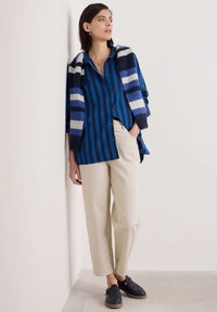 Camicia a righe blu con bottoni, scialle in lana a righe blu e bianche, pantaloni beige e scarpe slip-on nere intrecciate.