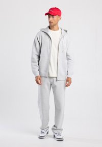 Hombre con una gorra roja, camiseta blanca, sudadera con cremallera gris claro, pantalones de chándal a juego y zapatillas blancas, de pie frente a un fondo neutro.