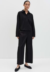 Vrouw die staat in een loszittend zwart knoopshirt, wijde zwarte broek en zwarte flats tegen een effen lichte achtergrond.