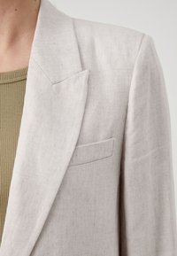 Blazer in lino grigio chiaro con reverse a punta, taschino sul petto singolo, texture morbida e vestibilità rilassata. Indossato sopra un top in verde oliva con texture.