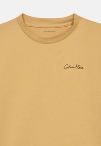 Κοντομάνικη μουστάρδα μπλούζα φτιαγμένη από μαλακό βαμβάκι, με στρογγυλή λαιμόκοψη και μικρό μαύρο λογότυπο "Calvin Klein" στο στήθος.