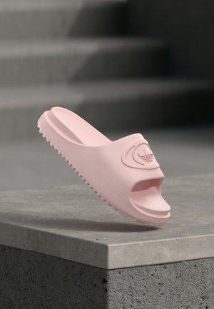 Hellrosa Gummisandale mit geriffelter Sohle und geprägt großem Logo, positioniert auf einer Betonmauer mit Treppen im Hintergrund.