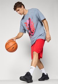Blå grafisk t-shirt med pink og sort design; røde træningsshorts; sorte basketballsko; hvide sokker med sort logo; basketball i hånden.