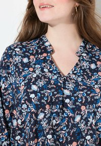 Marineblauwe bloemenblouse met korte mouwen, voorzien van een V-hals en kleine plooien; versierd met blauwe, roze en oranje bloemen tegen een donkere achtergrond.
