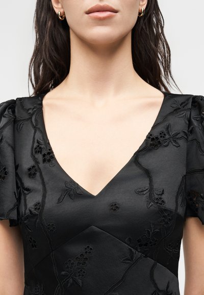 Γυναίκα που φοράει μαύρο φόρεμα με V-λαιμόκοψη, κεντήματα με λουλούδια και φουσκωτά μανίκια, συνοδευόμενο από χρυσούς κρίκους σκουλαρίκια.