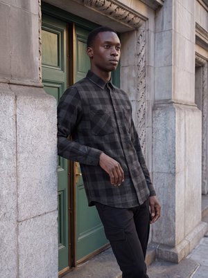Jeune homme portant une chemise à carreaux foncés et un pantalon noir, appuyé contre un bâtiment en pierre avec des portes à panneaux verts.