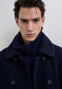 Mørkeblå dobbelbrystet frakke med stor struktureret krave, fremstillet af blødt, fluffy materiale. Under frakken ses en ribbet mørkeblå sweater.