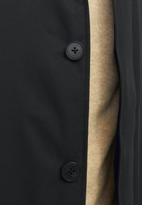 Tejido de abrigo negro con textura suave, presenta un botón negro prominente. Una prenda de color beige claro, texturizada, es parcialmente visible por debajo.
