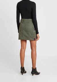 Jupe en denim olive verte, mini, avec deux poches arrière et des bandes sur les côtés en rouge, bleu et blanc. Portée avec un haut noir ajusté à manches longues.