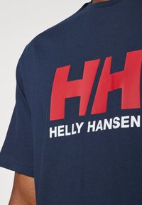 Tamno plava pamučna majica s velikim crvenim i bijelim logotipima "HH" i "HELLEY HANSEN" na prednjoj strani. Dizajn s okruglim izrezom, kratki rukavi.