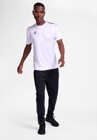 Camiseta atlética blanca con cuello en V y detalles en negro, combinada con pantalones jogger negros que presentan un patrón de chevrones en el lateral y un logo.