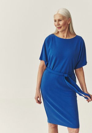 Starsza kobieta z długimi siwymi włosami, ubrana w niebieską sukienkę do kolan z paskiem w talii, spogląda nieco w prawo i się uśmiecha.