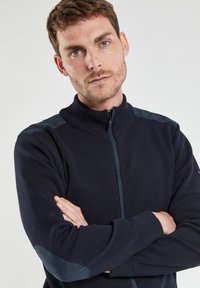 Sweat-shirt zippé marine avec un col côtelé, un corps texturé et des accents : épaule lisses. Comprend une fermeture éclair sur toute la longueur à l'avant et des poignets ajustés.