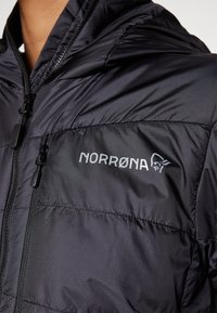 Svart isolert jakke med glidelås foran. Den har et glatt, skinnende stoff og "NORRØNA"-logo i hvitt på brystlommeområdet.