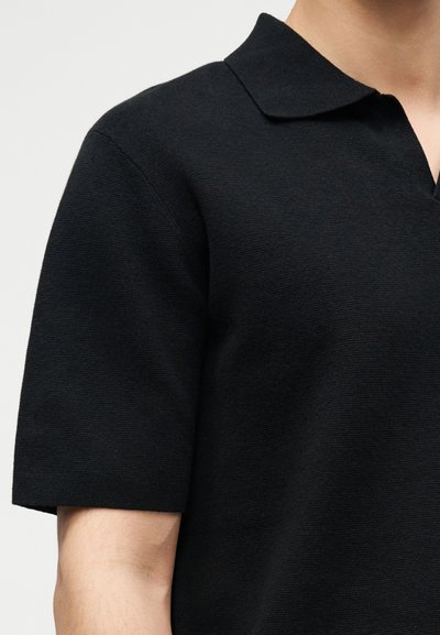 Μαύρο πλεκτό πόλο με κοντά μανίκια που φοριέται από άτομο με ανοιχτόχρωμο δέρμα, με ορατό γιακά, απλό φόντο.