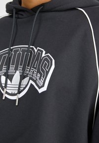 Czarna bluza z kapturem z dużym białym logo Adidas na piersi, ozdobiona srebrnymi akcentami na sznurkach oraz kontrastującymi białymi lamówkami po bokach.