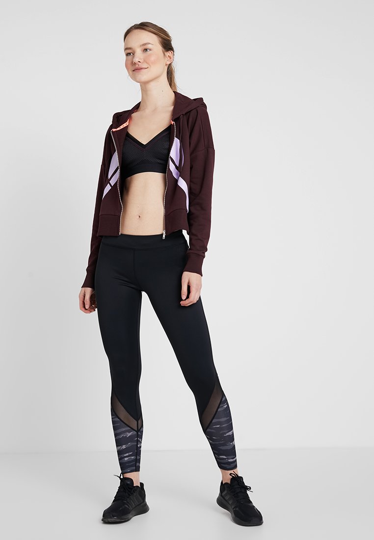Bordeauxkleurige hoodie met ritssluiting en paarse accenten, zwarte sportbeha en zwarte leggings met een textuurpatroon en netpanelen; gedragen met zwarte sneakers.