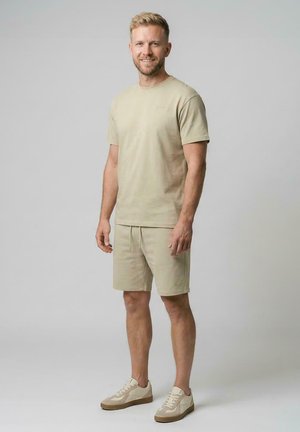 Glimlachende man staand, gekleed in een beige kortemouwenhemd, bijpassende korte broek en lichtgekleurde sneakers tegen een effen achtergrond.
