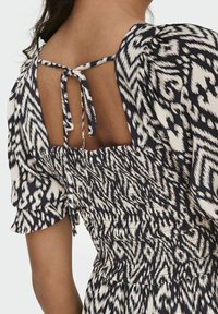 Robe présentant un motif ikat noir et crème, une taille smockée et des manches bouffantes. Le dos possède une découpe avec une fermeture à lien.