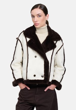 Donna che indossa un cappotto in shearling bianco e marrone scuro sopra un dolcevita beige con le mani nelle tasche dei pantaloni scuri.