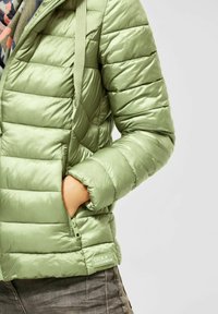 Grøn quilted jakke med hånd i lynlåslommen, båret over trykt skjorte og grå bukser, med "Cecil Outerwear" mærket.