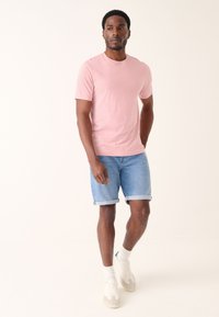 Lyserød bomulds T-shirt med korte ærmer kombineret med lyseblå denimshorts. Modellen bærer hvide sneakers med grå detaljer og hvide mellemlange sokker.