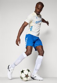 Hvid fodboldtrøje med blå accenter og logo, blå shorts, hvide sokker og sort-hvide støvler, placeret over en mønstret fodbold.