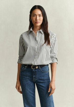 Frau mit langen dunklen Haaren, die ein weißes Hemd mit schwarzen vertikalen Streifen und blaue Jeans trägt, steht vor einem schlichten hellen Hintergrund.