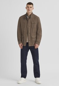 Braune, leichte Jacke mit vier Fronttaschen, Knopfverschlüssen und einem erhöhten Kragen. Getragen über einem grauen Shirt und blauen Jeans mit weißen Sneakers.