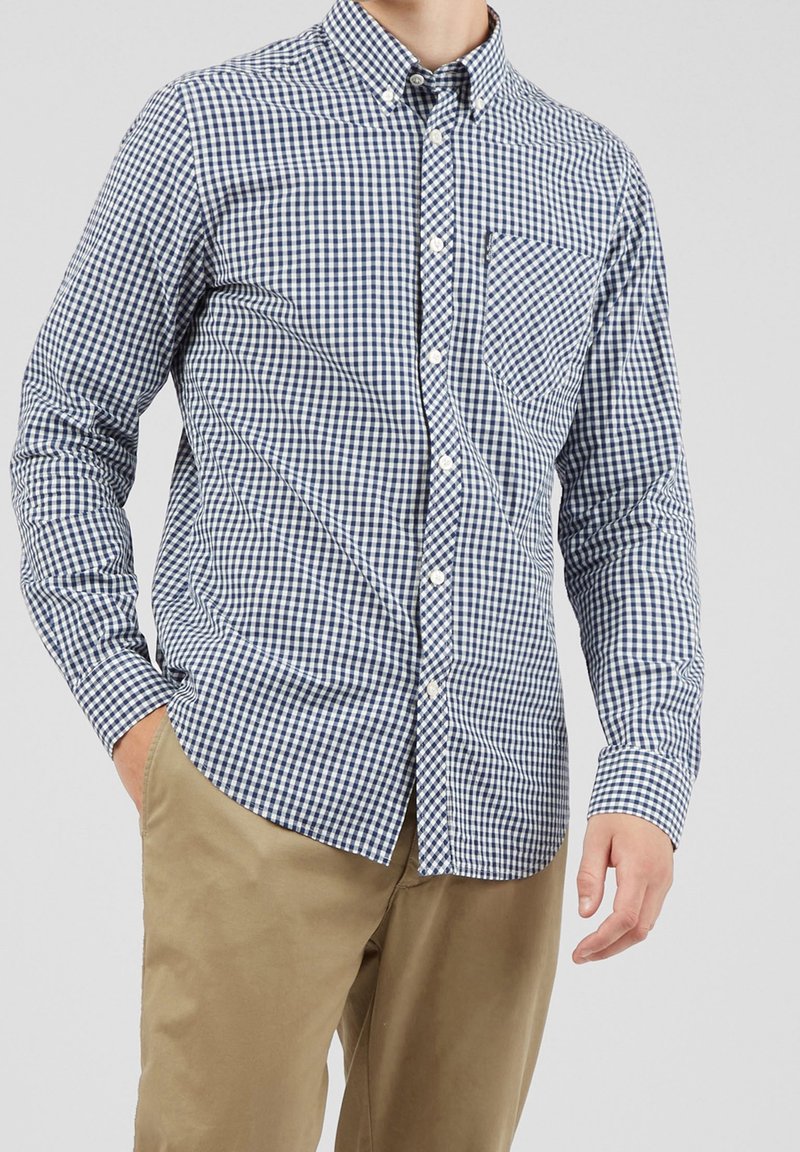 Blue and white checkered button-up shirt with long sleeves, a front pocket, and a classic collar, paired with khaki pants.