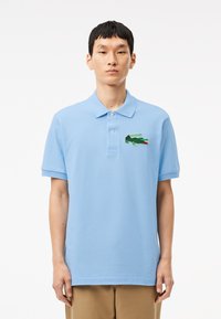 Ljusblå polo skjorta i bomull med korta ärmar, krage och en broderad grön krokodil-logotyp på vänster bröst.