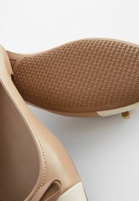 Ruskea liukas kenkiä, jossa on sileä päällinen, vaaleita beige-värisiä yksityiskohtia ja teksturoidut kumipohjat, joissa on timanttikuvio. Mukana on pieni kultainen yksityiskohta.