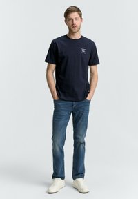 T-shirt di cotone blu navy, maniche corte, scollo rotondo, con testo bianco sottile sul petto; abbinato a jeans azzurri e sneakers bianche.