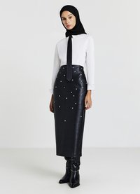 Jupe en cuir noir ornée de perles, complétée par une chemise blanche et une cravate noire. Le modèle porte des bottes noires montantes jusqu'aux genoux.