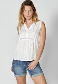 Blusa bianca senza maniche con dettagli ricamati e scollatura a V, abbinata a pantaloni corti in denim blu con accenti floreali.