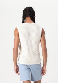 Strikket pullover i creme med diamantmønster, ærmeløs design og en tætsiddende pasform, parret med lyseblå shorts.