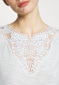 Vit topp med intrikata spetsdetaljer vid halsringningen, tillverkad av mjukt tyg med en subtil struktur. Spetsen har blommiga och geometriska mönster.