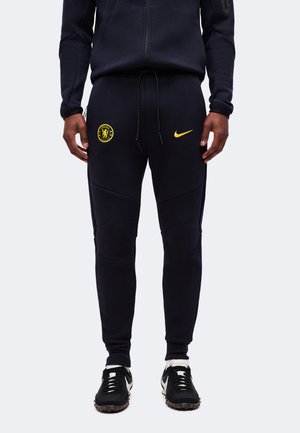 Personne portant un pantalon de jogging noir avec le logo jaune de Chelsea FC et le swoosh jaune de Nike, associé à des baskets noires et blanches.