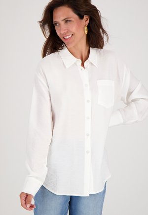 Vrouw die glimlacht en een witte blouse met knopen en een voorzak draagt, met een lichtblauwe spijkerbroek, poserend met één hand op haar heup.
