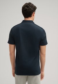 Marineblaues Poloshirt mit klassischem Kragen, kurzen Rollärmeln und glatter Textur. Verfügt über Nähdetails an den Schultern.