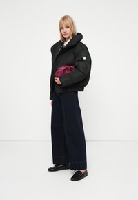 Jachetă puffer neagră cu guler înalt, purtată cu pantaloni din denim întunecat cu croială largă. Ținând o posetă maronie, cu mocasini negri care accentuează ținuta.